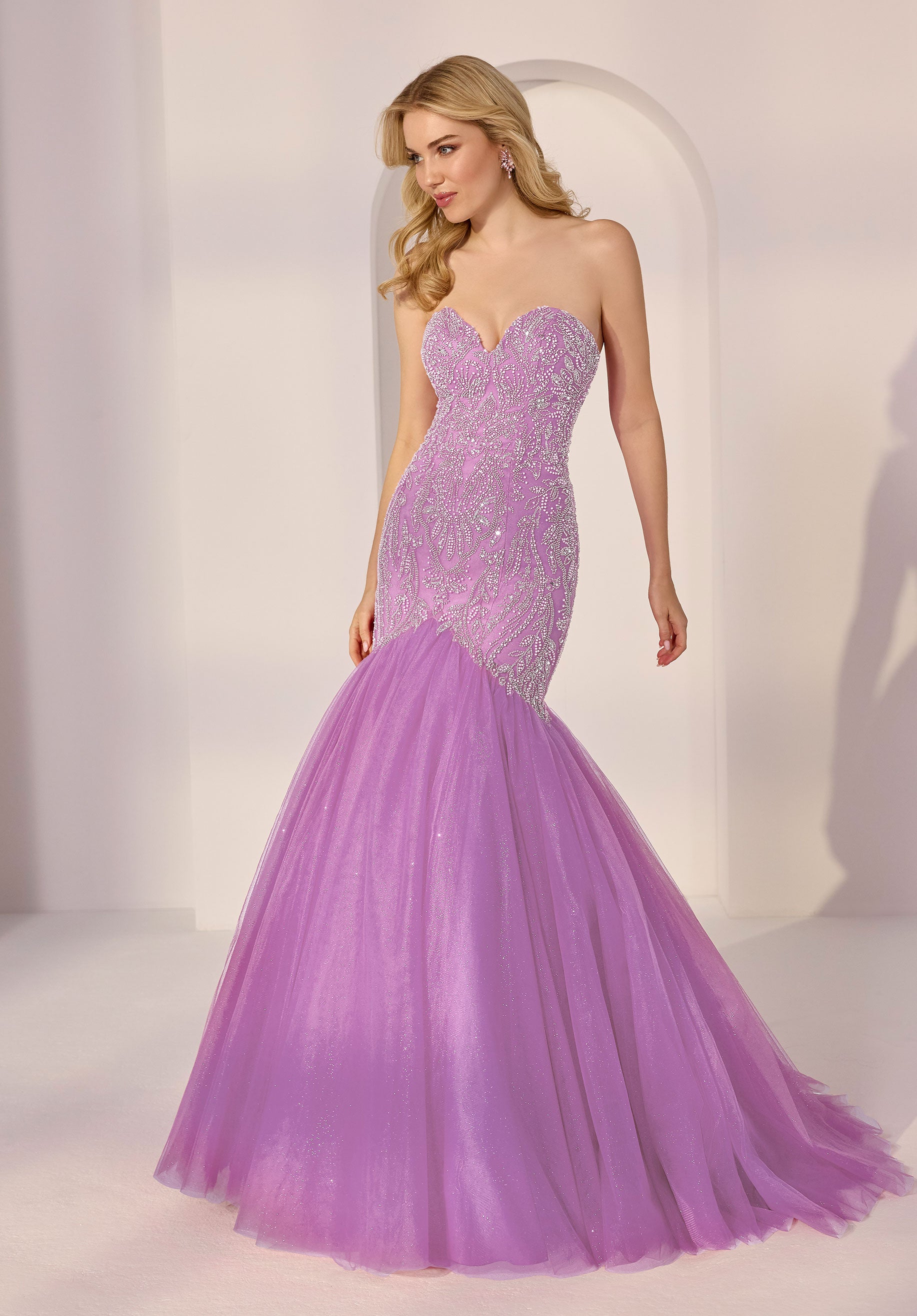A woman in a lavender Morilee 49203 strapless mermaid prom dress with a sequin bodice and tulle skirt stands in a softly lit room.
